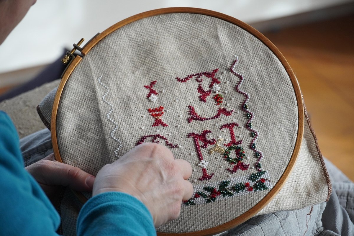 Person som broderar ett rött och grönt julbroderi med bokstäver och motiv i träring på grått linne