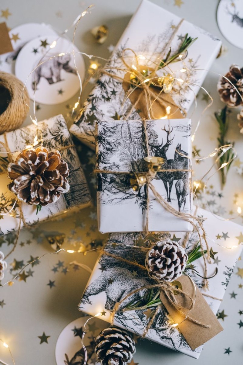 Svartvita paket med hjortmotiv dekorerade med kottar, rosmarin, snöre, kraftetiketter och ljusslinga, med guldstjärnor