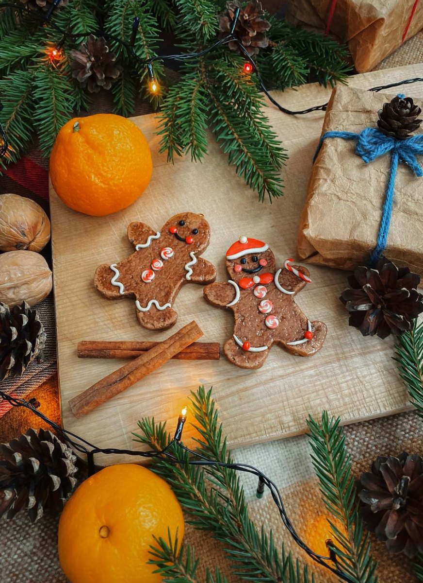 Två pepparkaksgubbar med vit glasyr och röd-vita detaljer, en med tomteluva, på träbräda med kanelstänger, mandariner och granris